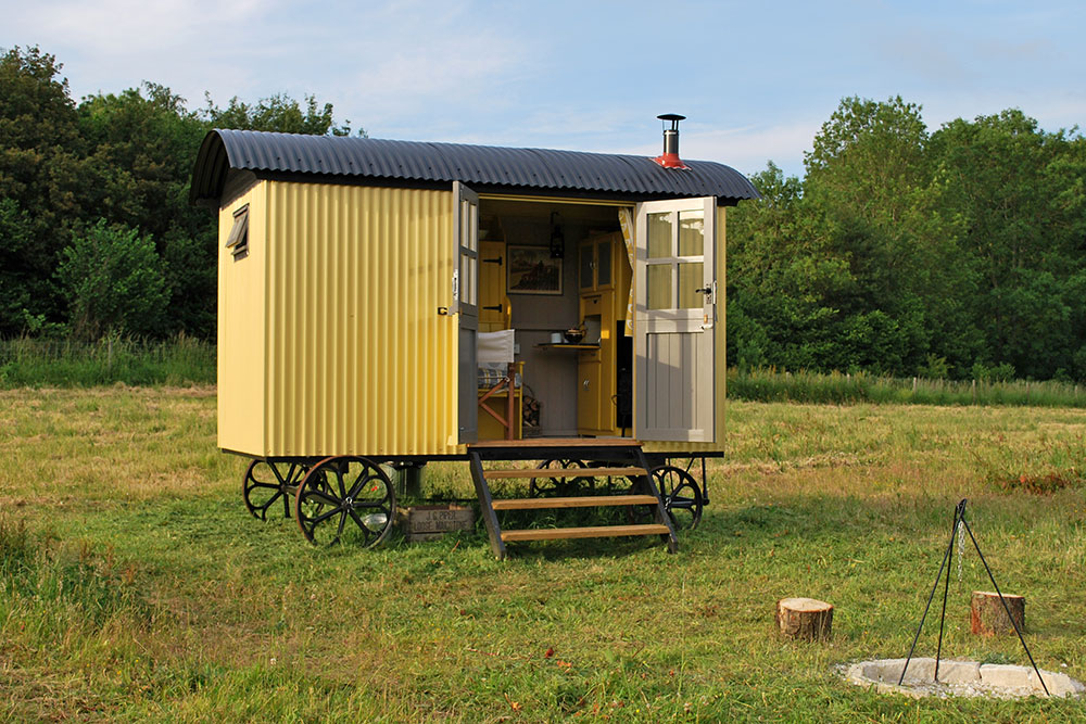 For a shepherds hut holiday in Hampshire, Barrow shepherd's hut is perfect with open double doors to the interior, campfire and log seating in the foreground. Find out about Glamping at Wallops Wood