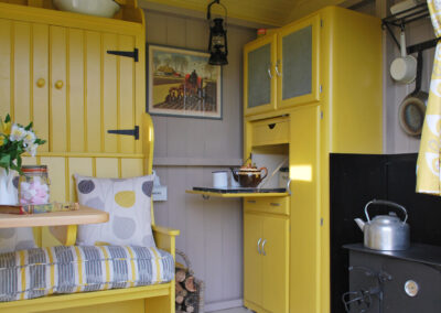 Cosy shepherds hut glamping in the living area in Barrow shepherds hut with bench seat, storage cupboards, table with jar of marshmallows, hurricane lamp hanging from the ceiling, woodburning stove with kettle on the top, and saucepans hanging on the wall.