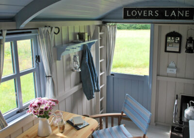 Beautifully decorated Beacon shepherd's hut interior in shades of ivory and pale blue with painted wood-panelled walls, stable door open to the meadow, jar of marshmallows and books on the table with director's chair, and woodburning stove with kettle on the top. For guest information, click the links on this page