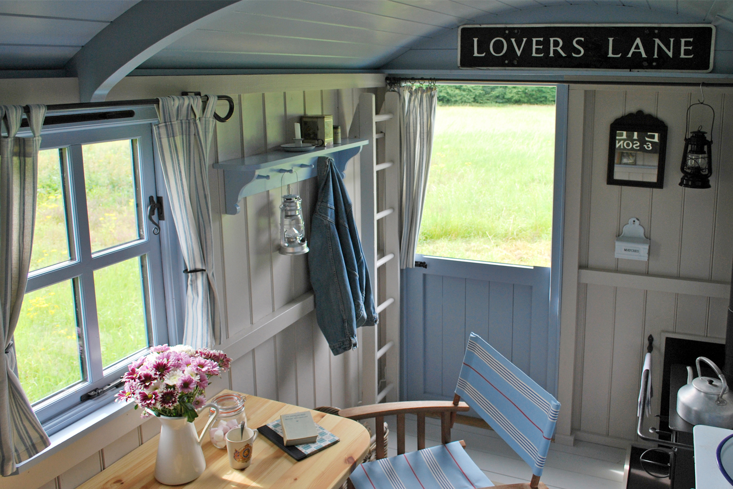 Beautifully decorated Beacon shepherd's hut interior in shades of ivory and pale blue with painted wood-panelled walls, stable door open to the meadow, jar of marshmallows and books on the table with director's chair, and woodburning stove with kettle on the top.<br />
For guest information, click the links on this page