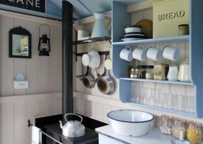 The kitchen space in Beacon shepherd's hut with woodburning stove and kettle on top, enamel bowl, saucepans hanging on the wall and enamel crockery and glassware on the shelving