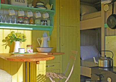 Boundary shepherd's hut interior showing table and chairs, wash stand, shelving for crockery, woodburning stove with kettle, cooking pans hanging on the wall and two single beds