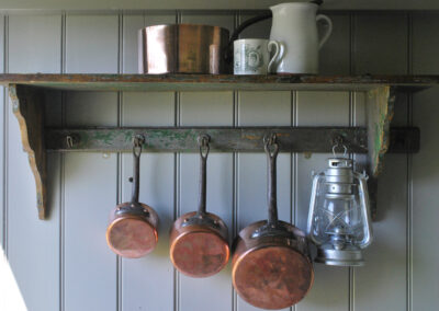 Copper pans and an oil-fuelled hurricane lamp hanging from shelving hooks with white jug, large copper pan and mug on top of the shelf