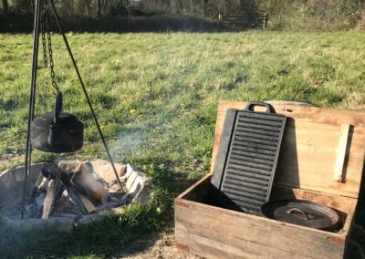 Lit campfire with large black kettle hanging from a cooking tripod with open box next to it containing a cooking griddle and large pan