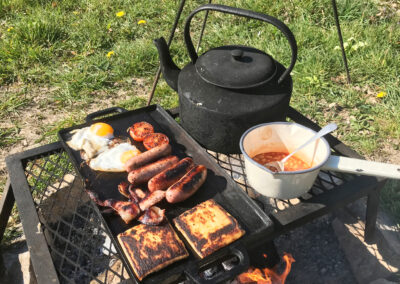 Large black kettle on the trivet, pan of baked beans and eggs, bacon, sausages, fried bread and tomatoes being cooked over the firepit