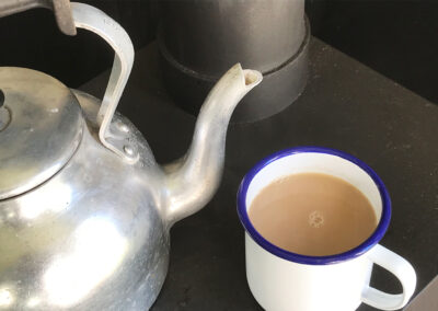 Stainless steel large kettle next to a hot mug of tea in a white enamel mug on top of the woodburning stove