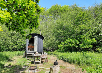 Old Winchester on a glorious sunny day with deep blue sky and surrounded by lush green vegetation and ancient woodland with stable doors open to the interior, picnic table and director's chairs and campfire with large black kettle and log seating in front