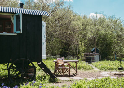 Open windows and stable door of Old Winchester looking out onto the paddock with picnic table and director's chairs, hot tub, and campfire, surrounded by woodland on a sunny day with blue sky. What could be better on a shepherds hut holiday?