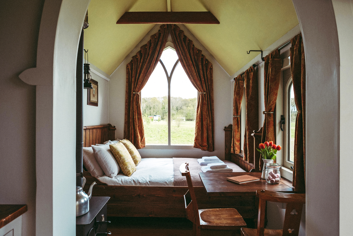 Interior of St Brides replica chapel tiny glamping house with double bed, arched ceiling and windows looking out onto the meadow. Enjoy this space during your shepherds hut holidays in Hampshire
