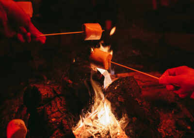 Two marshmallows on sticks being held over the lit campfire and giving a warm red glow