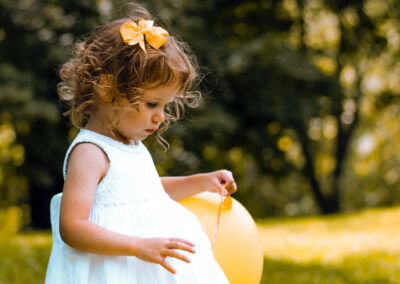 Female toddler with dark curly hair wearing a white summer dress and holding a yellow balloon with the same colour bow in her hair playing on the meadow and enjoying our child friendly holidays
