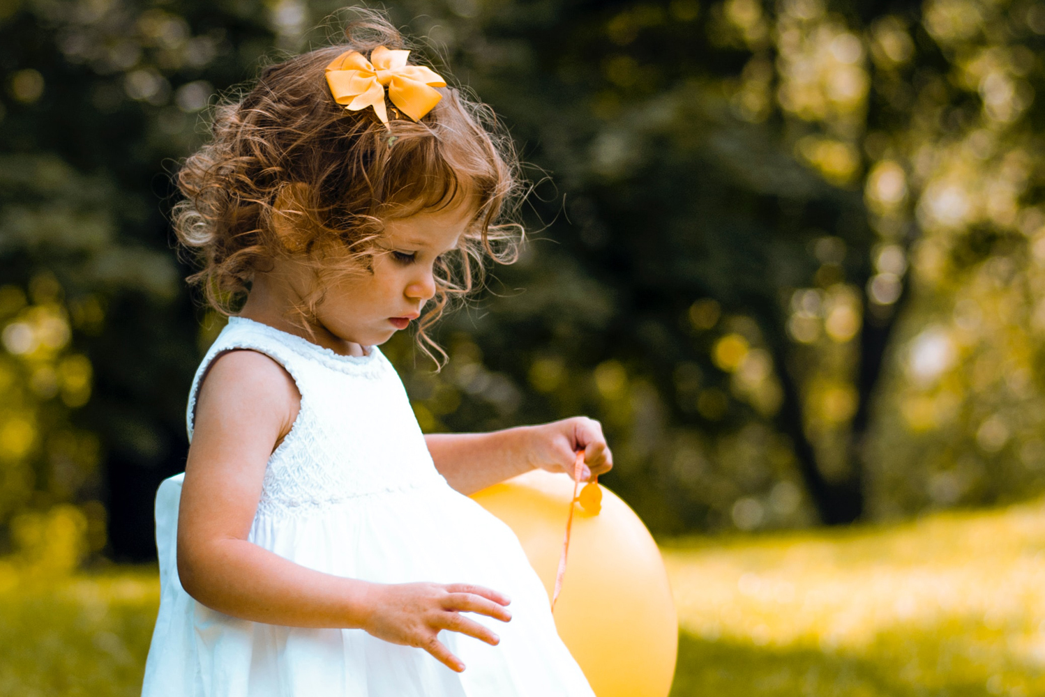 Female toddler with dark curly hair wearing a white summer dress and holding a yellow balloon with the same colour bow in her hair playing on the meadow