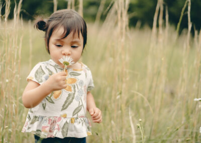 Dark-haired toddler, wearing a flowering top and dark trousers, smelling a daisy in her hand as she walks through the long grass at Wallops Wood