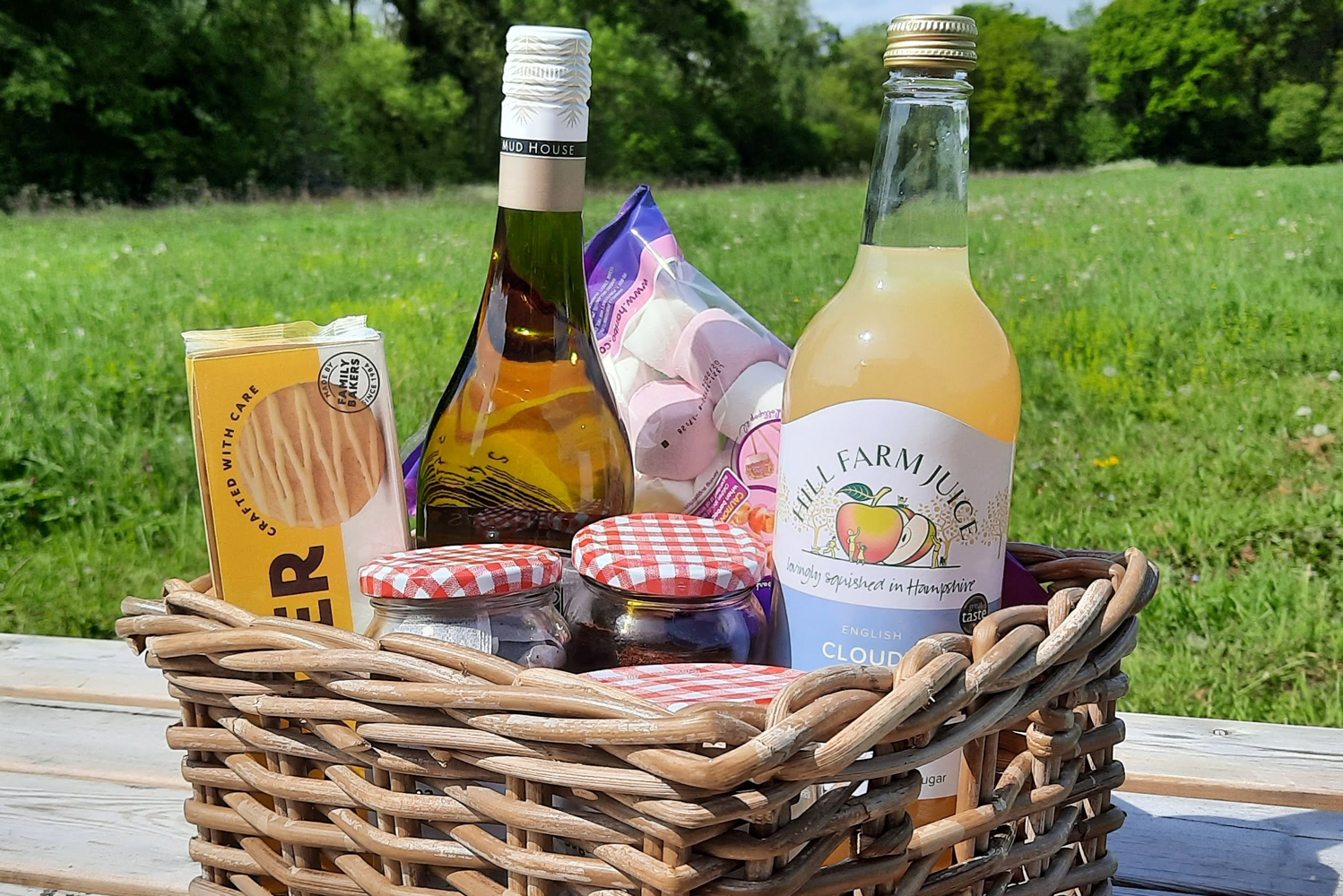 Our Welcome Hamper in a wicker basket on a picnic table surrounded by green grass on a sunny day containing tea, coffee, sugar, locally produced apple juice, wine, marshmallows and biscuits is what to expect in your Shepherd's Hut