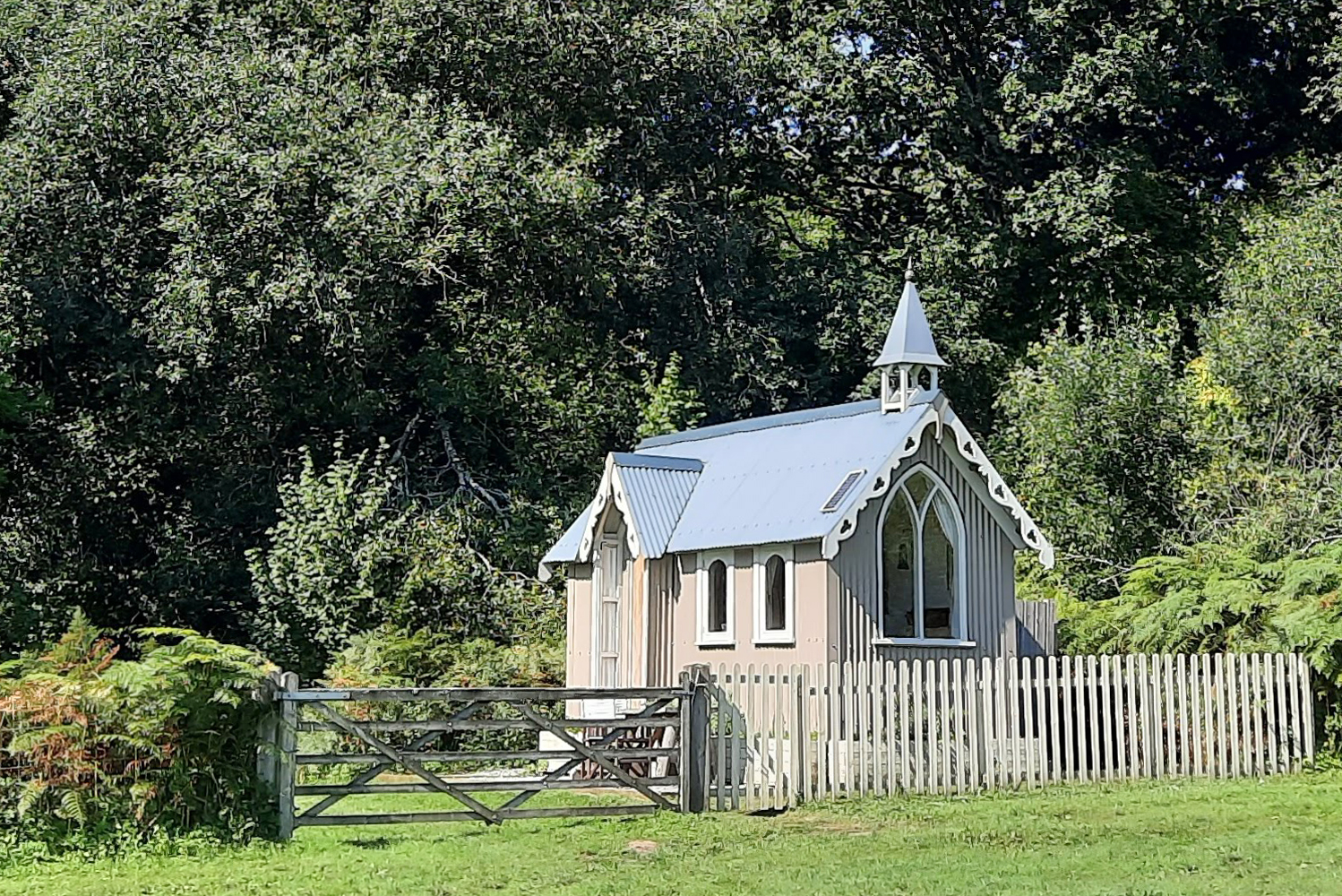 St Bride's shepherds hut with ensuite on a sunny summer day from the meadow showing the five bar gate and picket fence into the garden with ancient woodland behind it